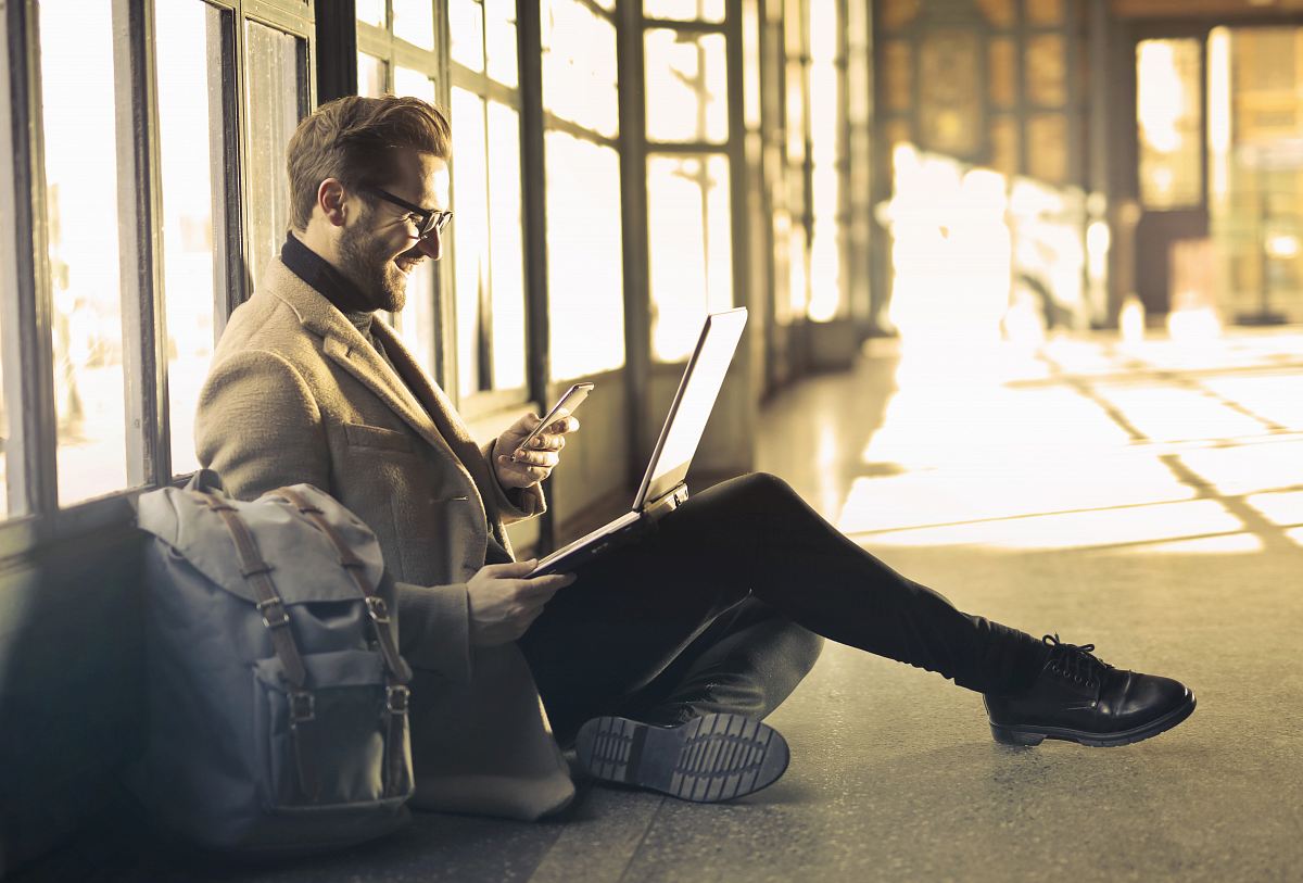 man sitting near window holding phone and laptop