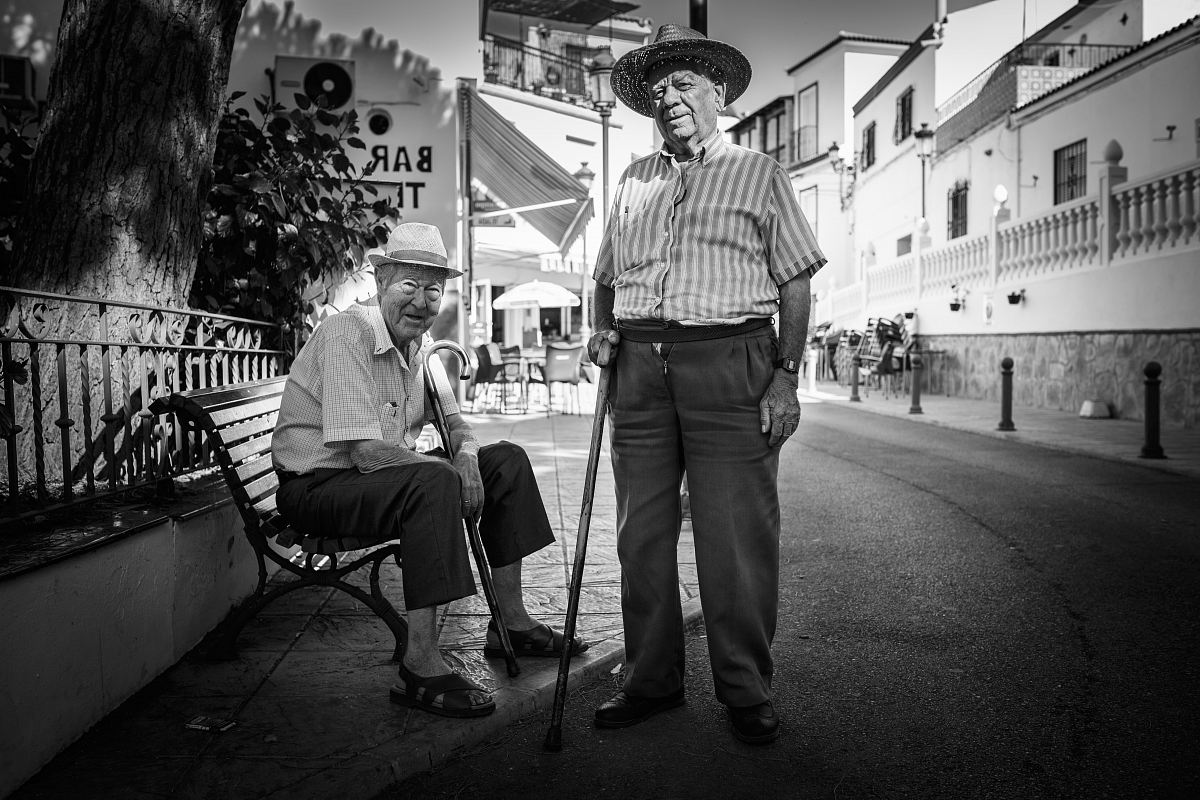 man standing beside a man sitting on cast iron bench near buildings