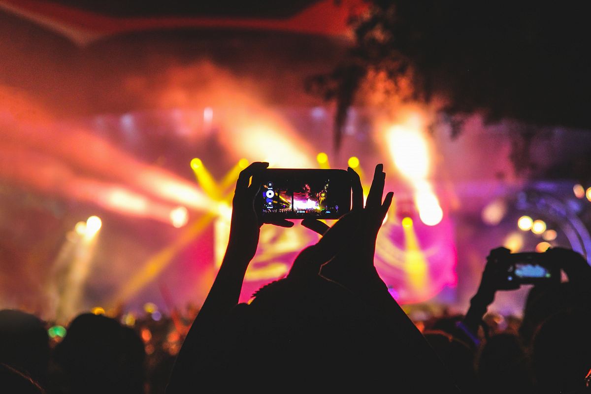 person holding smartphone taking video of a concert near stage with lights during nighttime