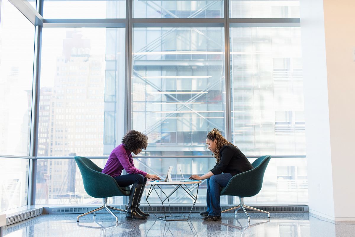 two women sits of padded chairs while using laptop computers