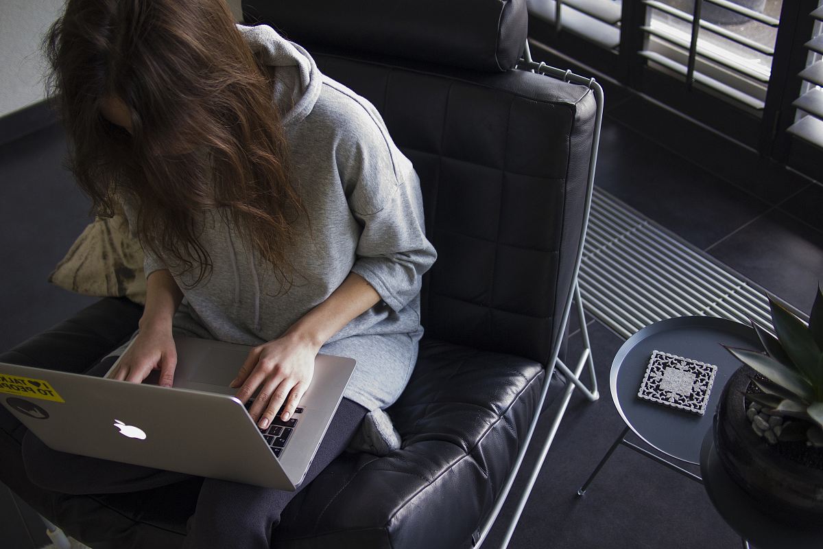 woman in blue chambray long-sleeved top sitting on black leather chair with silver MacBook on lap
