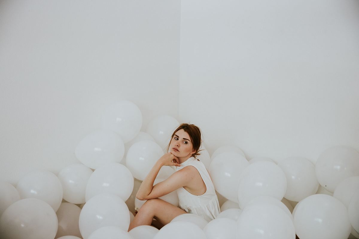 woman wearing white sleeveless tops sitting on floor surrounded by white balloons inside white painted room