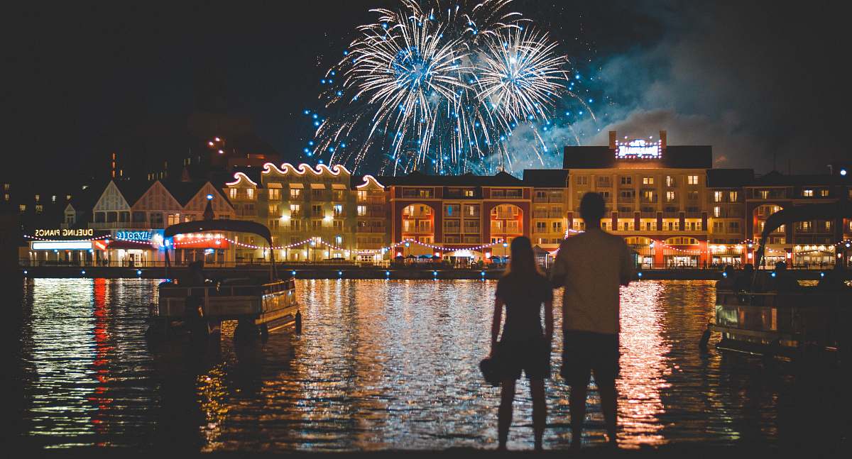  two people standing beside body of water watching fireworks 