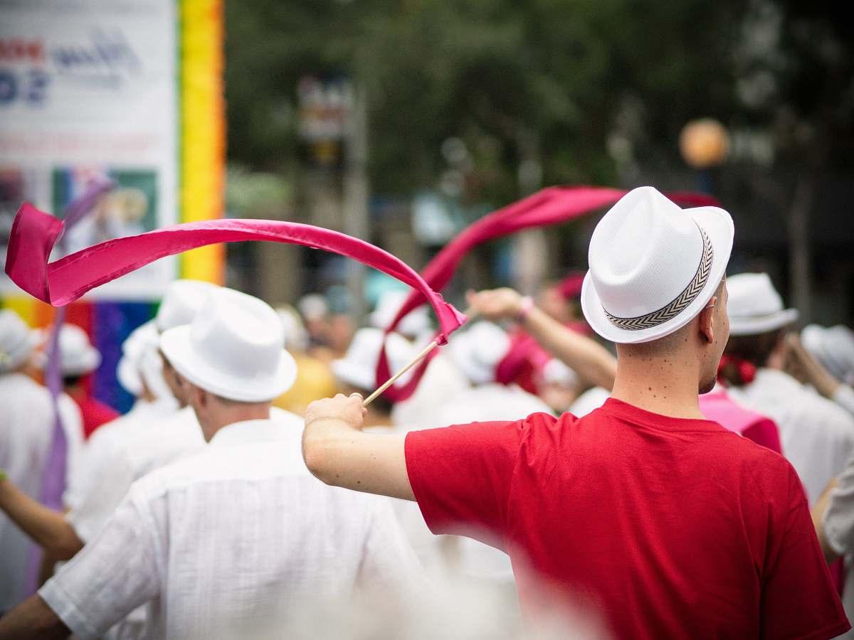 human selective focus photography of man wearing pink t-shirt and white fedora hat holding pink ribbon with stick waving on the air during daytime west hollywood