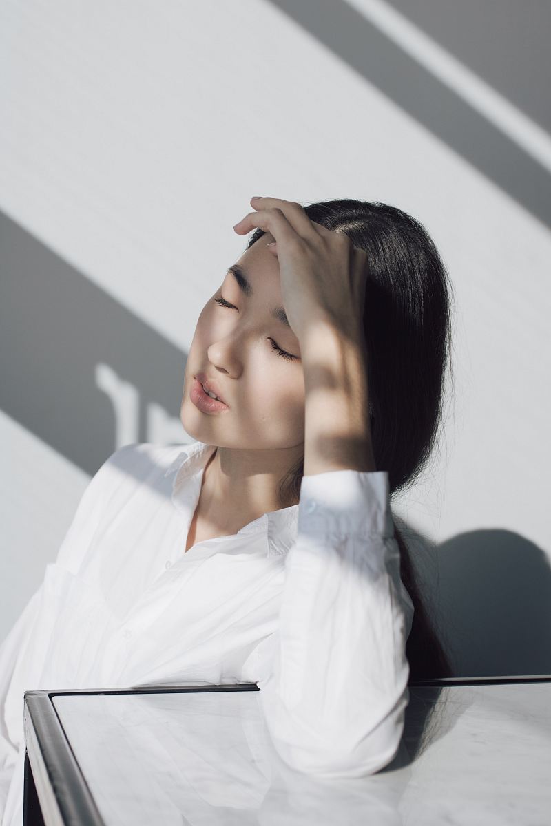 This poignant image captures a woman seated at a pristine white table, with her head resting in her hands, conveying a deep sense of contemplation and introspection. The serene, minimalistic environment highlights her emotional state, making it a perfect visual for themes of stress, reflection, or decision-making. Ideal for use in articles, mental health resources, or any creative project seeking to evoke feelings of vulnerability and thoughtfulness.