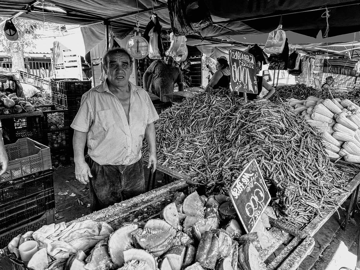 person grayscale photo of man in button up shirt standing in front of vegetables market
