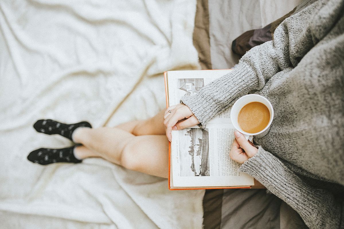 woman holding a cup of coffee at right hand and reading book on her lap while holding it open with her left hand in a well-lit room