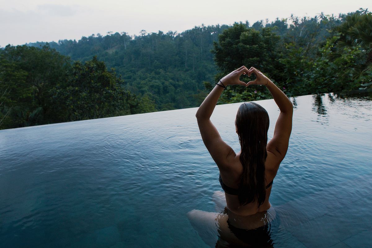 woman in infinity pool making heart hand gesture facing green leafed trees