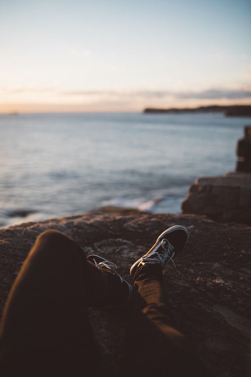 person wearing black pants and low-top sneakers lying down on brown rock formation near seashore