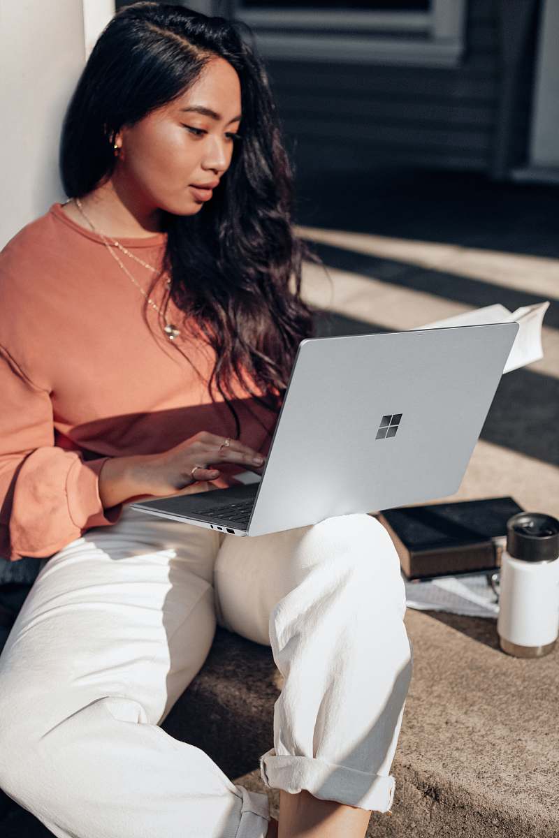 electronics woman in orange long sleeve shirt and white pants sitting on floor using Surface Laptop  pc