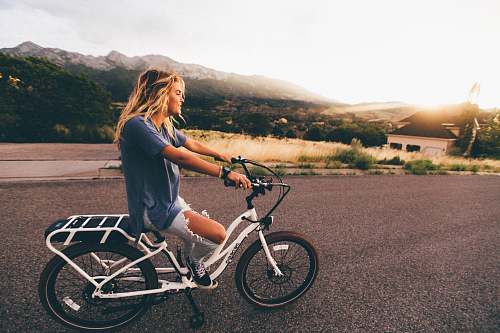 person woman in blue t-shirt and distressed blue denim jeans riding white bicycle on grey road near mountains at daytime human