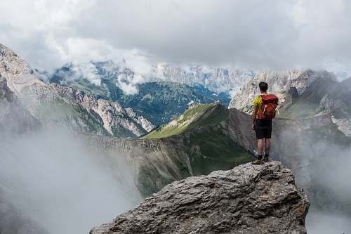 nature man with red backpack standing on cliff facing mountains under white sky during daytime hiking