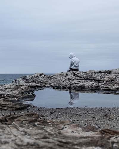 human man sitting on cliff overlooking sea during daytime puddle