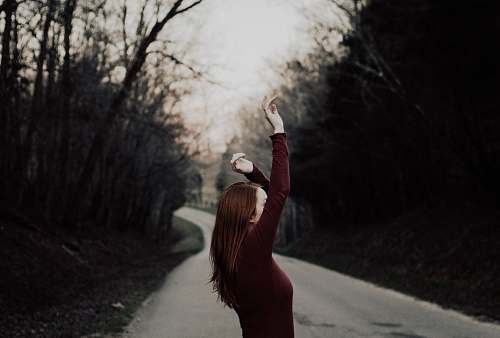 woman selective focus photography of female raising hands in the middle of the road surrounded by withered trees dirt road