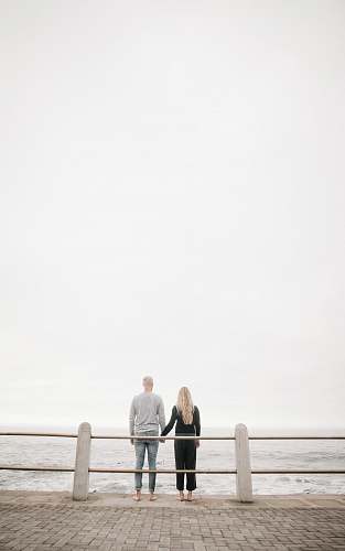 people couple leaning on guard rail while holdings hands facing ocean at daytime person