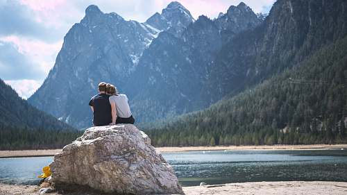 people man and woman sitting on rock near body of water and mountain during daytime couple