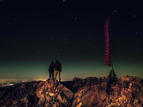 night silhouette of two people standing on brown rocks under starry night stars