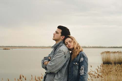 human woman in blue denim jacket leaning on man's shoulder near body of water people