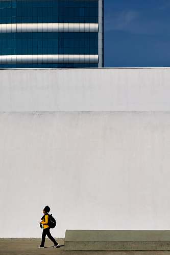 people human in yellow long-sleeved T-shirt walking beside white concrete wall human
