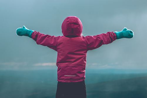 person wearing pink hooded jacket raising her hand in front of green mountain range during daytime