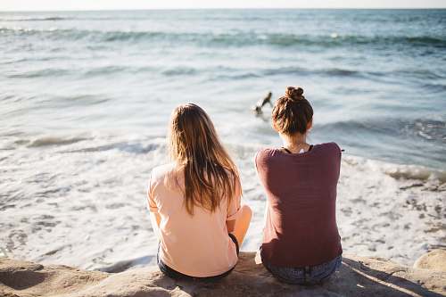 human two women sitting on cliff looking at the ocean beach