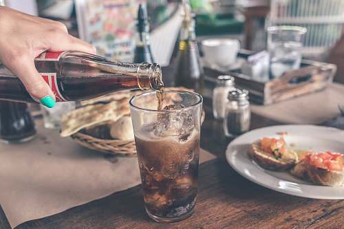beverage person pouring Coca-Cola soda on clear drinking glass near round white ceramic plate glass