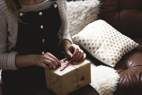 woman in grey sweater and blue denim dungarees sitting on brown and white sofa opening a yellow gift box closeup photography