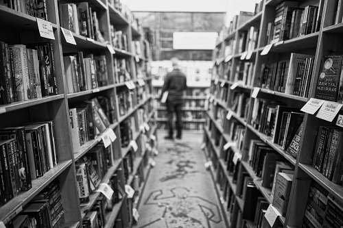 person grayscale photo of man standing in front of books black-and-white
