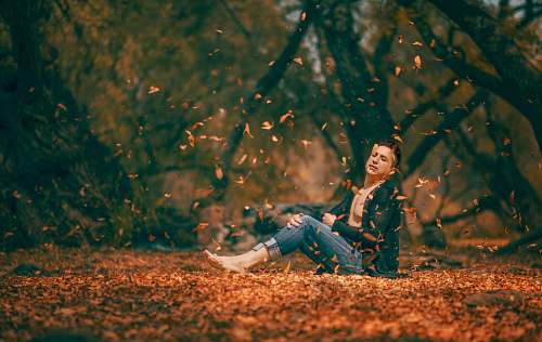 human shallow focus photography of man sitting on brown dried leaves surrounded by trees during daytime people