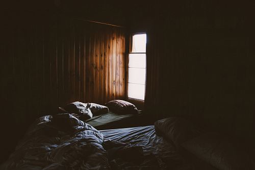 Pale light pouring into a cabin through a small window illuminates two unmade beds