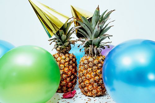 two pineapples with gold party hats near colorful balloons