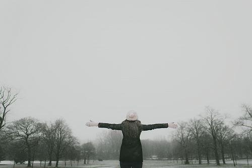 woman in black long-sleeved shirt standing and facing trees with stretched arms under white sky