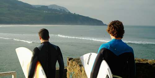 water two male wearing wetsuits holding surfboards standing nearby each other on top of hill with view of beach below surfboard