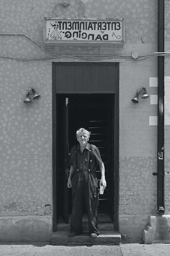 person grayscale photo of man wearing collared tops and dress pants standing between concrete wall near Entertainment Dancing signage building black-and-white