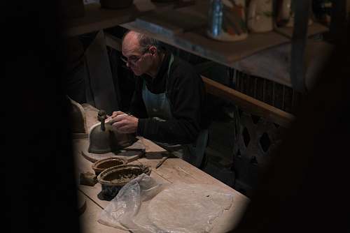 person man in black long-sleeved shirt and blue apron working on gray pot on table human