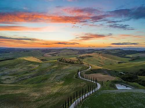 landscape road between green grass field near mountains under blue and brown sky at golden hour field