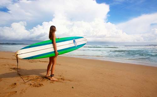 beach woman standing near sea holding white, blue, and green surfboard under blue sky coast