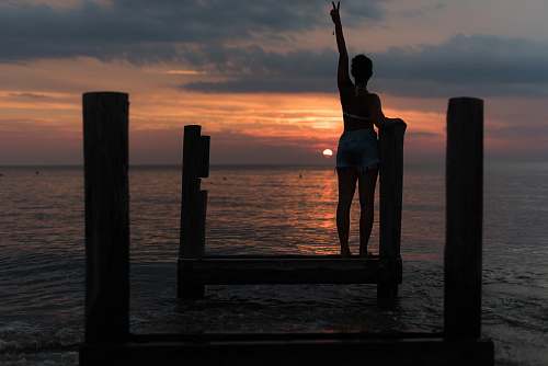 sunset woman standing on brown wooden dock rising her right hand during golden hour human