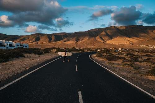 road person carrying surfboard on asphalt road near brown mountains surfer