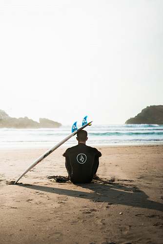 ocean man sitting on sand beside surfboard facing sea during daytime sea