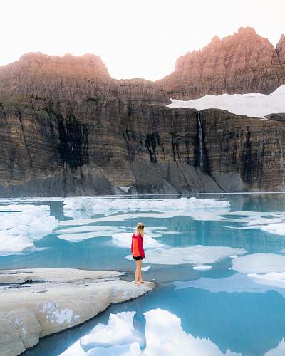 nature woman in red and orange sweater stands on stone beside water mountain