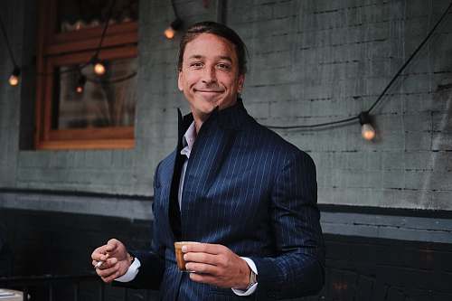 people man wearing black suit holding brown mug near black string lights human
