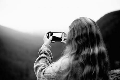 person grayscale photography of woman wearing sweater using smartphone taking picture black-and-white