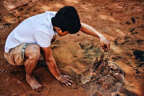 person Young man crouching on the beach drizzling sand and making sand castles mud
