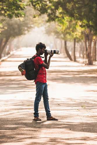 human man in red t-shirt holding black DSLR camera taking photos while standing on grey asphalt road during daytime apparel