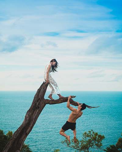 couple woman sitting on tree trunk with man holding on branch near sea under white clouds during daytime ocean
