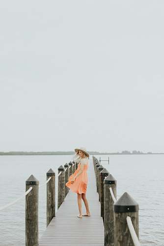 people woman in orange spaghetti strap mini dress standing on brown wooden dock during daytime woman