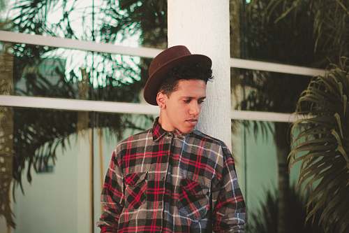 human man wearing brown fedora hat and plaid dress shirt while leaning on white post hat