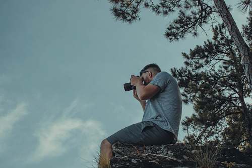 person man sitting on cliff while taking picture photography