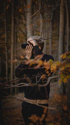 person photo of man wearing black jacket standing on forest taking photo photographer